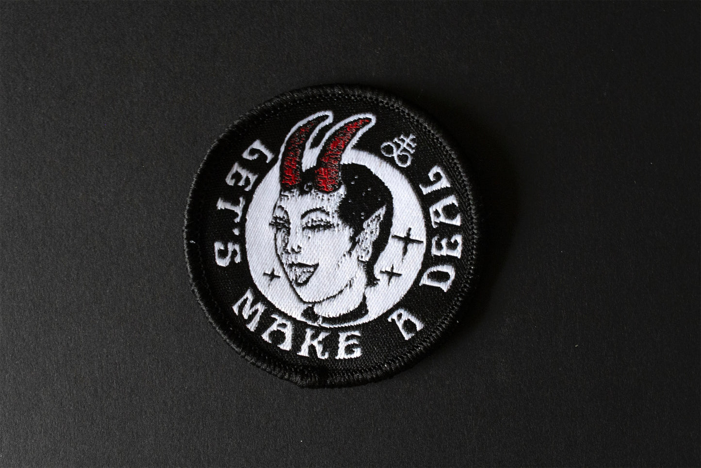 A round patch with the text let's make a deal, a devil woman winking, featuring a leviathan cross