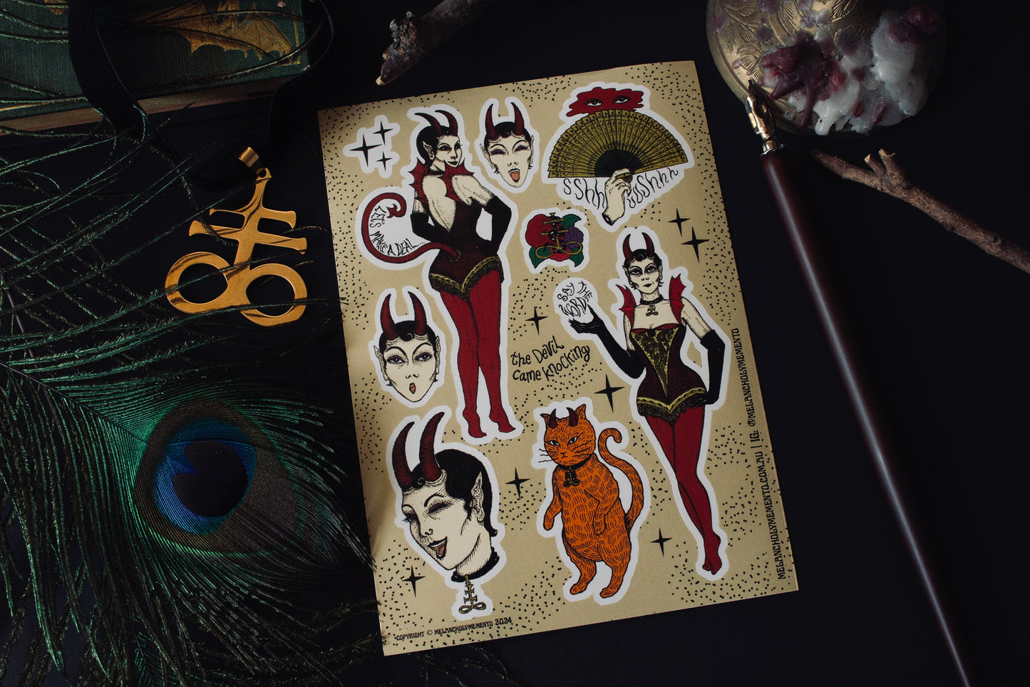Devil Came Knocking sticker sheet surrounded by peacock feathers, leviathan cross, and a calligraphy pen.