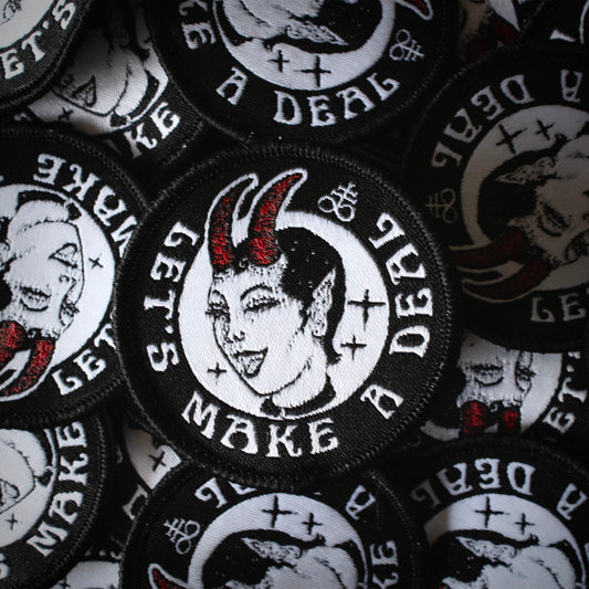 A round patch with the text let's make a deal, a devil woman winking, featuring a leviathan cross